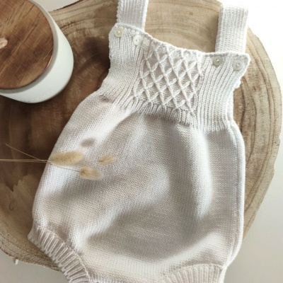 Macacão de bebé branco em malha sobre superfície de madeira com frasco branco ao lado.