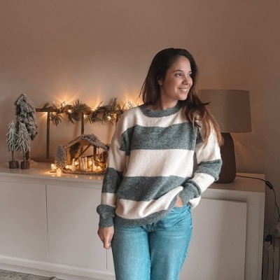 Pessoa vestindo camisola riscada, jeans azuis e sapatilhas brancas, em sala com decoração natalícia iluminada.