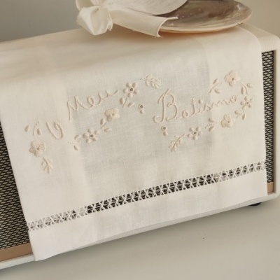 Toalha bege com bordado floral e texto 'O Meu Bolirno' sobre objeto retangular