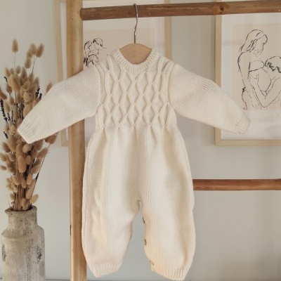 Macacão de bebé creme em malha grossa com padrão trançado pendurado em cabide de madeira
