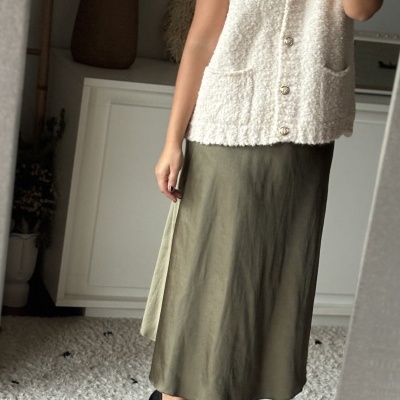 Mulher a usar colete creme e saia verde no interior com candeeiro e móvel ao fundo