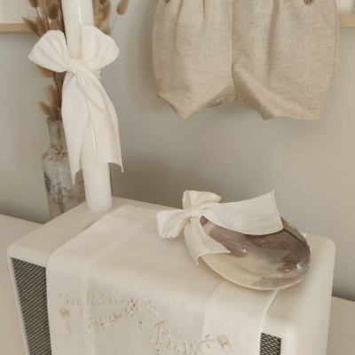 Velas decorativas com laços brancos, calças de bebé cinzentas, toalha branca bordada com texto e concha decorativa