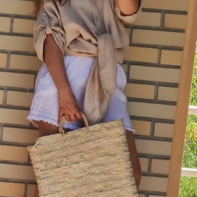 Mulher com roupa bege e branca segurando mala de palha em frente a parede de tijolos