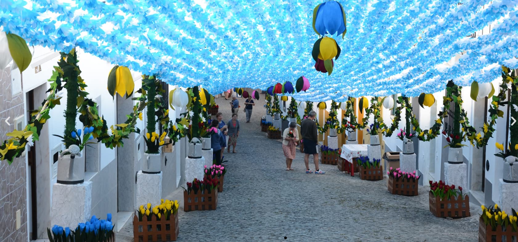 Festas do Povo de Campo Maior 2026 - Pack Hotel + Entrada Rua coberta por flores azuis artificiais suspensas e vasos com flores coloridas nas paredes e chão