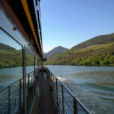 Passadiço de barco com corrimão metálico num rio entre colinas verdes sob céu azul