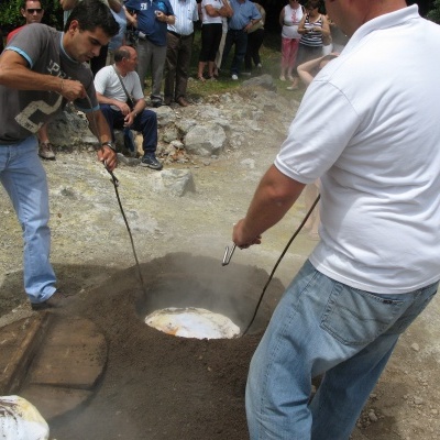 Homens a abrir forno subterrâneo em ambiente exterior rochoso com observadores ao fundo