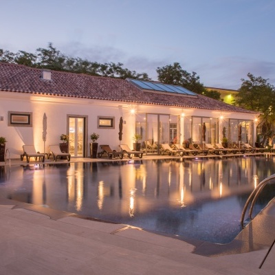 Piscina exterior com espreguiçadeiras e edifício iluminado ao entardecer