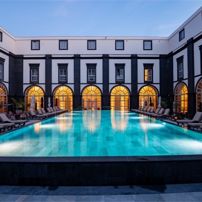 Piscina exterior iluminada num pátio de edifício com janelas arqueadas e espreguiçadeiras