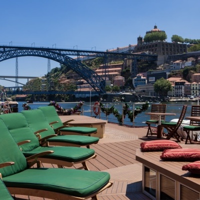 Espreguiçadeiras e cadeiras de madeira com almofadas verdes num convés de madeira com vista para a ponte Dom Luís I