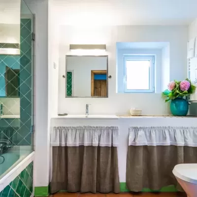 Casa de banho com banheira, lavatório, espelho, janela, toalheiros e vaso flores.