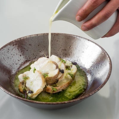 Prato de cerâmica com peixe, pão e molho verde, com molho claro sendo servido por uma mão