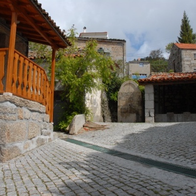 Área pavimentada com casa de pedra e varanda de madeira, fonte de pedra e edifício com telhado de telhas vermelhas