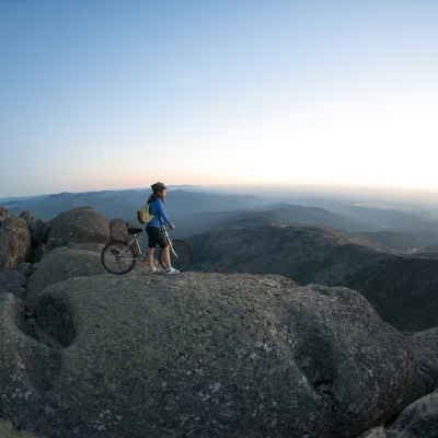 Pessoa com capacete e bicicleta sobre rocha com montanhas ao fundo.