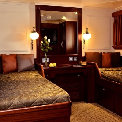 Quarto com duas camas de madeira escura, colchas castanhas, almofadas, espelho grande, candeeiros e cortinas castanhas.