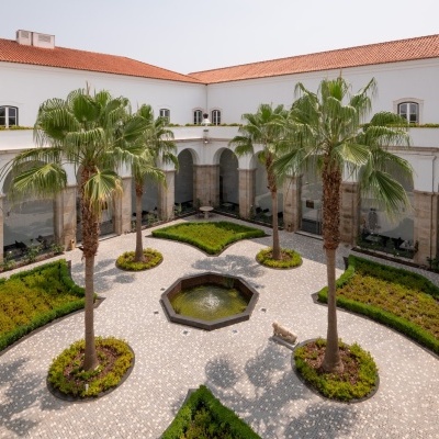 Pátio interior com palmeiras, fonte, canteiros e arcadas em edifício branco com telhado vermelho