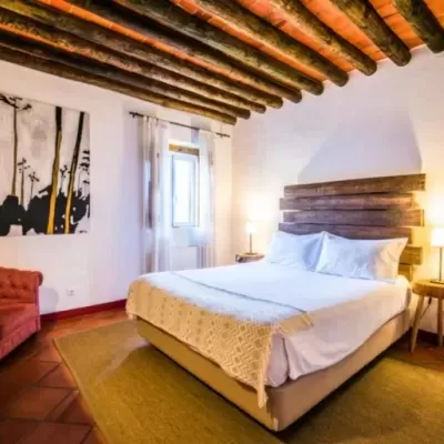 Quarto com cama de casal, cabeceira de madeira rústica, roupa branca, poltrona vermelha e teto com vigas de madeira