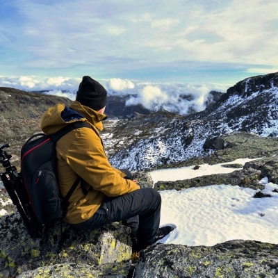 Pessoa sentada em rochas com casaco amarelo e mochila na natureza montanhosa com neve.