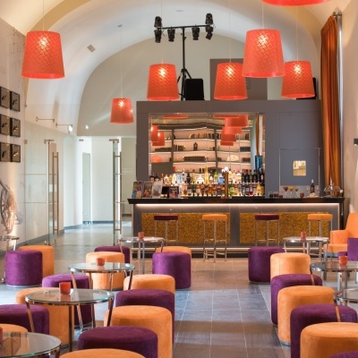 Sala de bar com bancos e pufes laranja e roxos, candeeiros pendentes laranja e balcão com azulejos dourados.