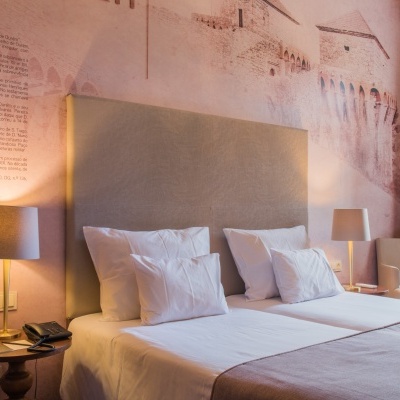 Quarto com duas camas individuais, cabeceira bege, roupa branca e manta rosa, mesas com candeeiros, poltrona e janela aberta.