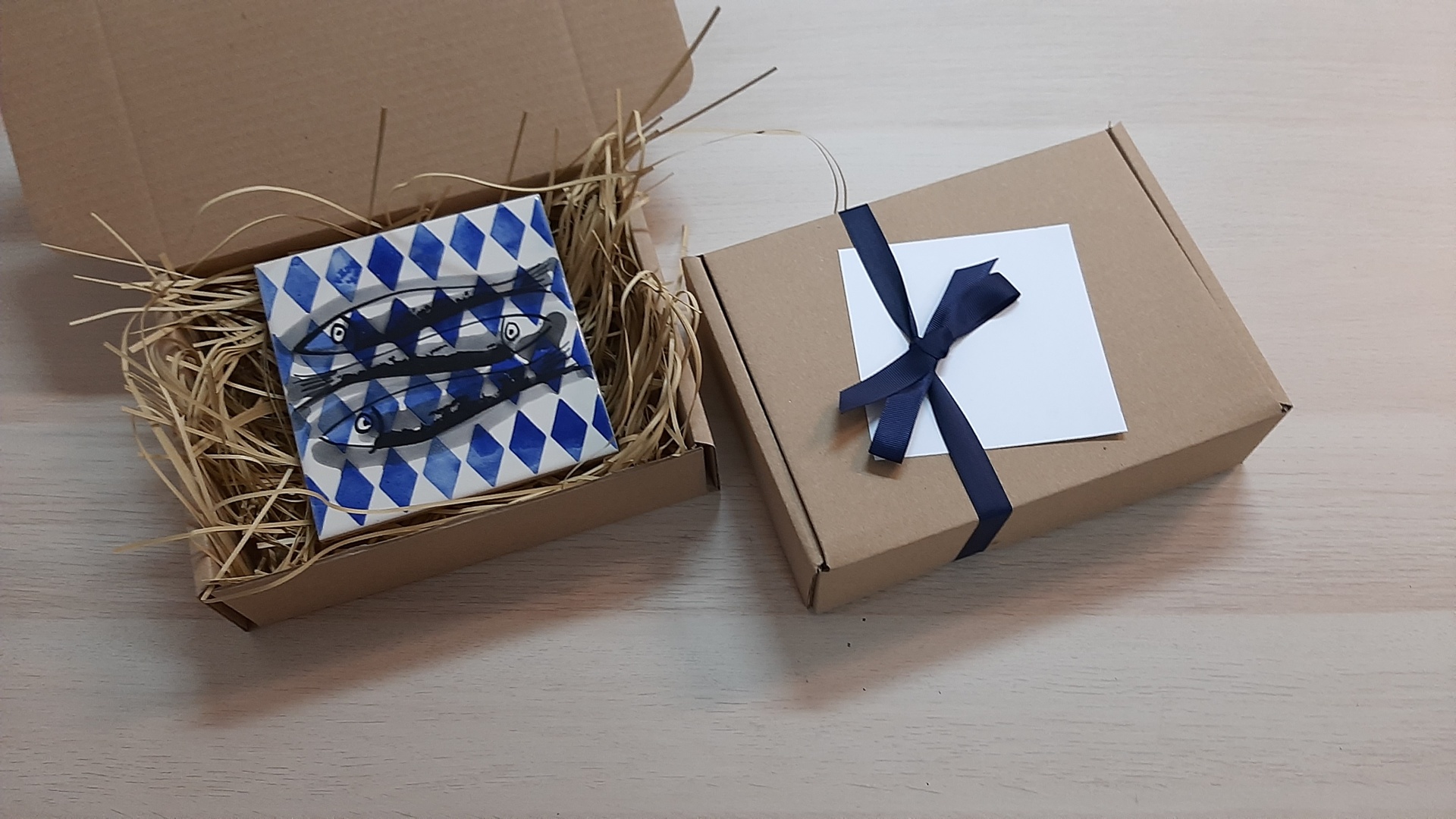 Two cardboard gift boxes, one open with a ceramic tile and straw, the other closed with a navy ribbon and white card
