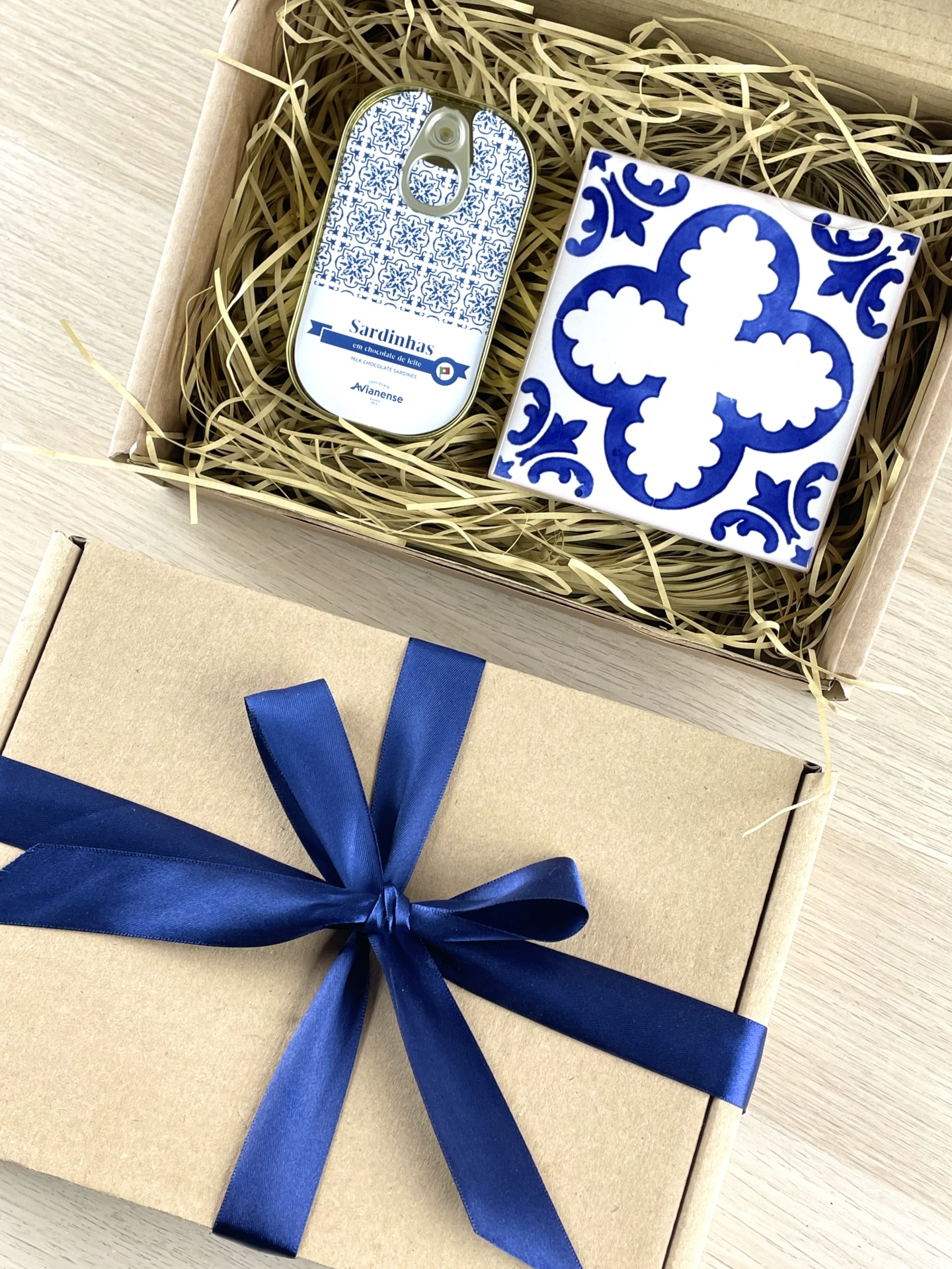 Gift box with tin of milk chocolate sardines and blue-white ceramic tile, open with straw filler and closed box tied with blue ribbon.