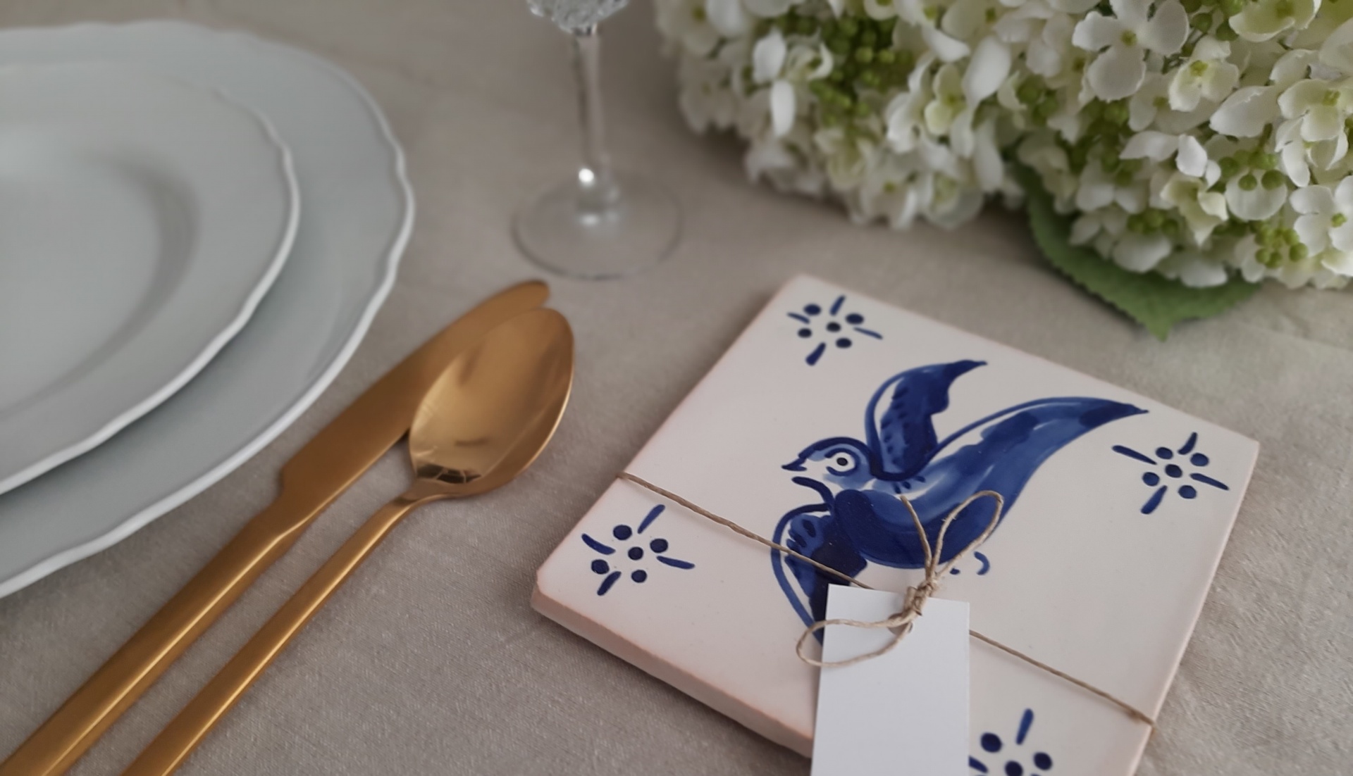 Ceramic tile with blue bird design tied with twine and tag on table with gold cutlery, white plates, glass, and flowers.