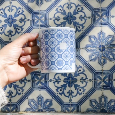 White ceramic mug with blue floral geometric pattern held against blue and white tiled wall