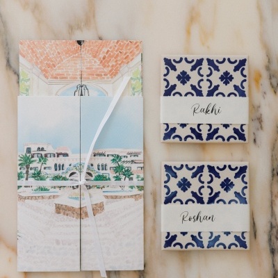 Two blue and white patterned boxes labeled Rakhi and Roshan next to a folded illustrated card tied with white ribbon on marble surface
