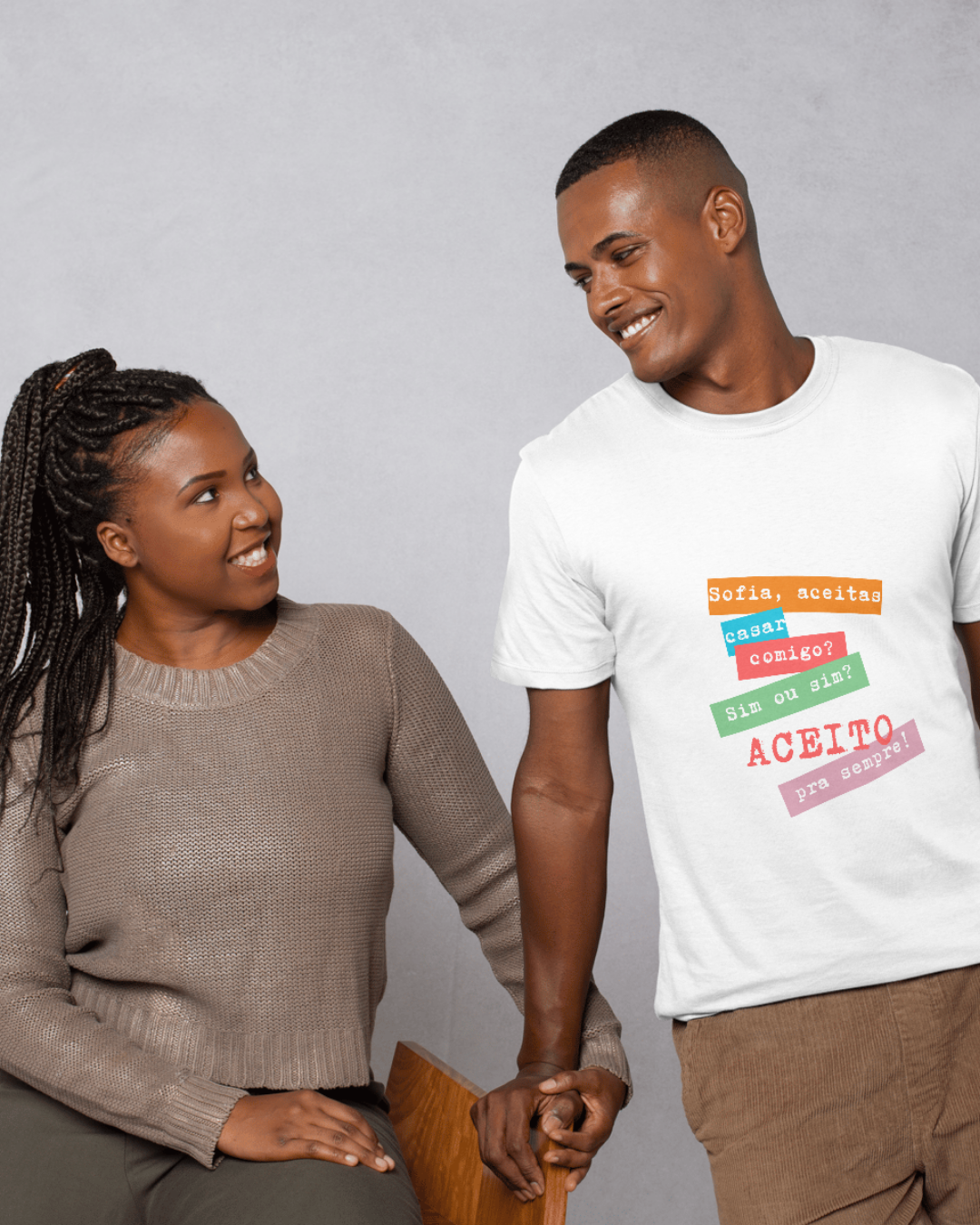 Casal sorridente de mãos dadas, homem com t-shirt branca com texto em português e mulher com camisola castanha.