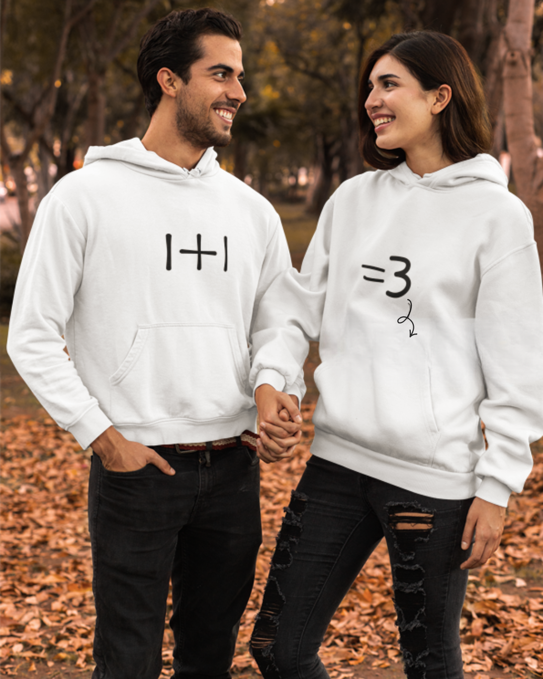Casal com sweatshirts brancas estampadas a sorrir numa área com folhas secas e árvores.