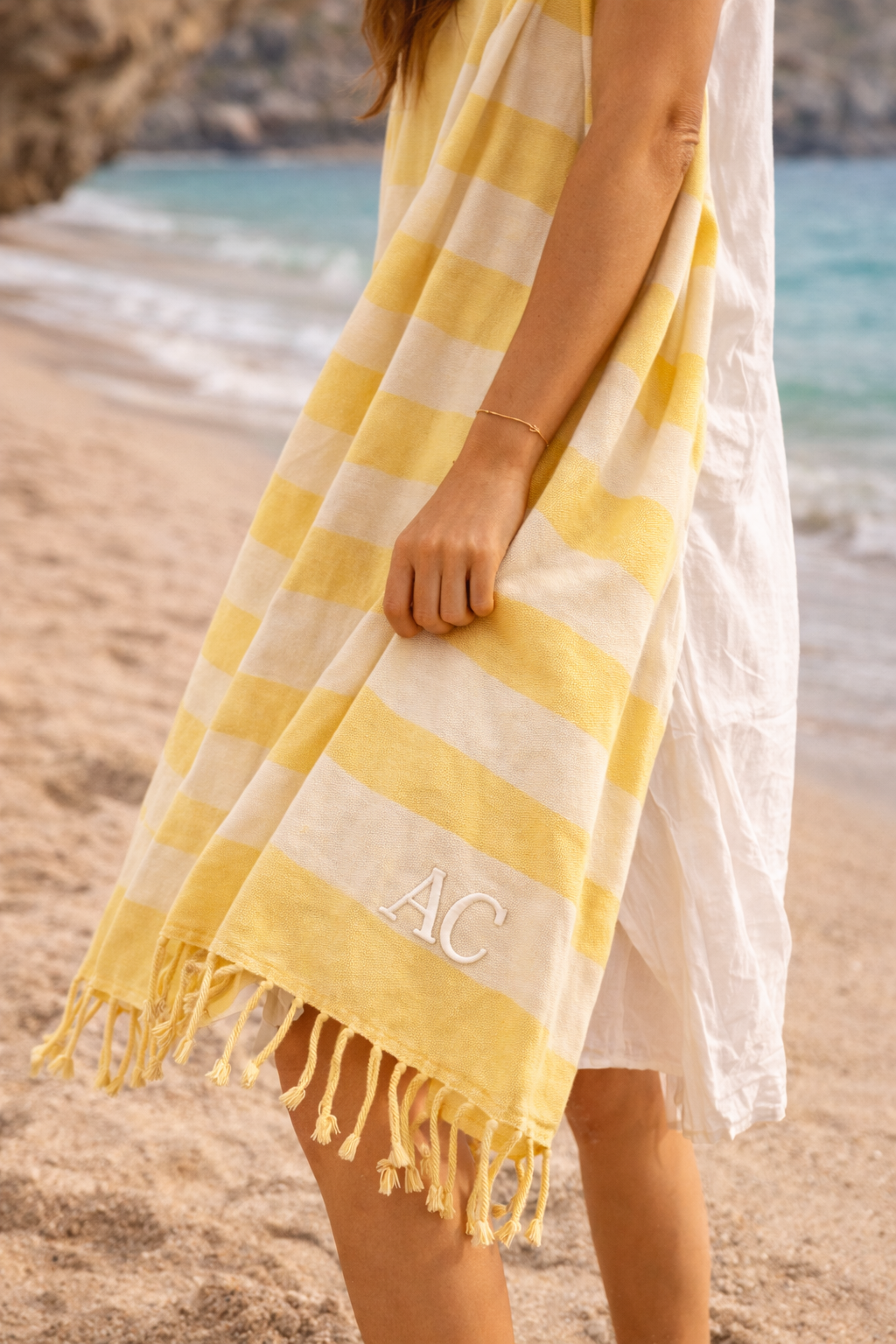 Manta de praia às riscas amarelas e brancas com franjas e monograma AC, segurada por pessoa junto ao mar.