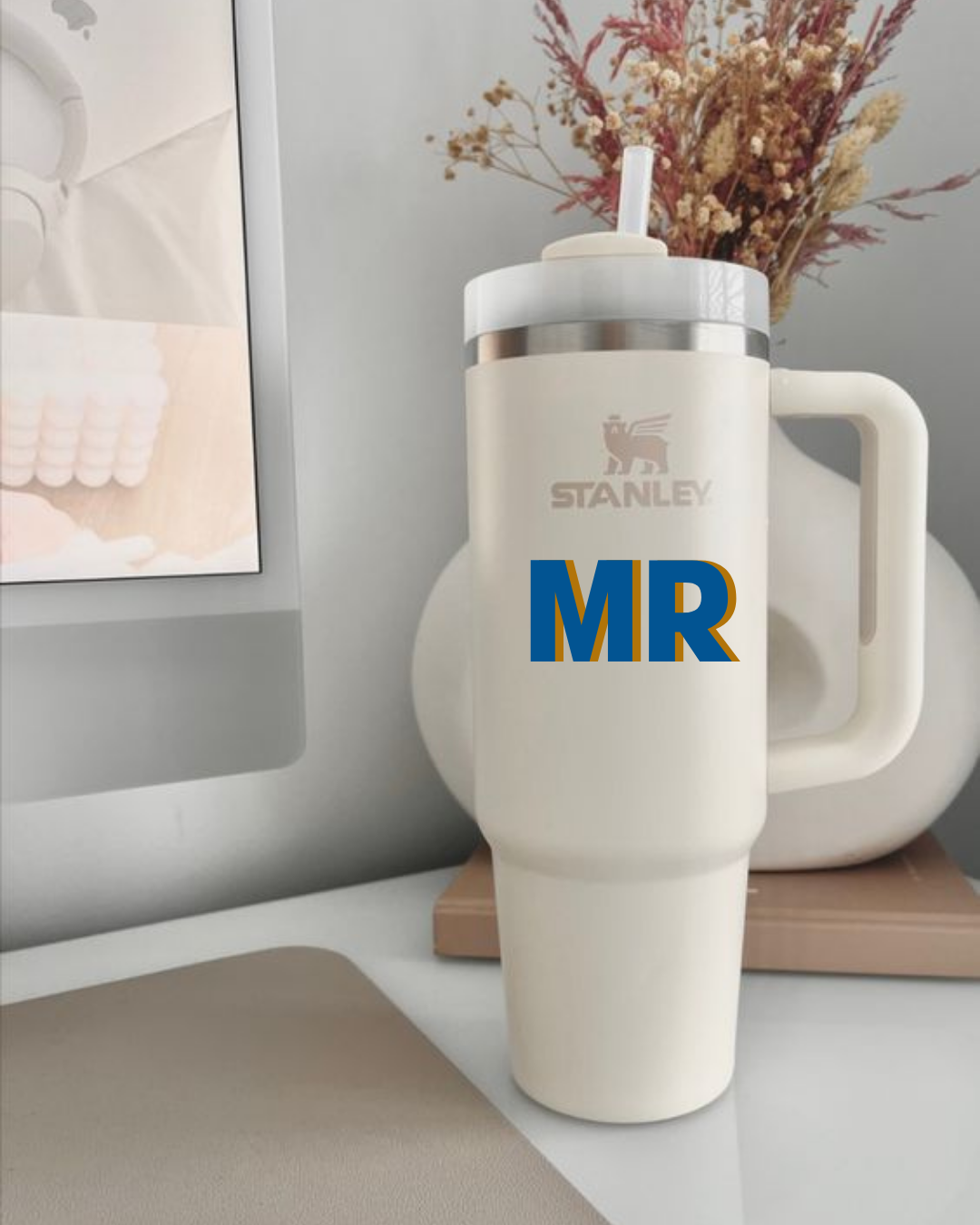 Copo térmico Stanley branco com alça grande e texto 'STANLEY' e 'MR' em azul e amarelo numa mesa com teclado e monitor