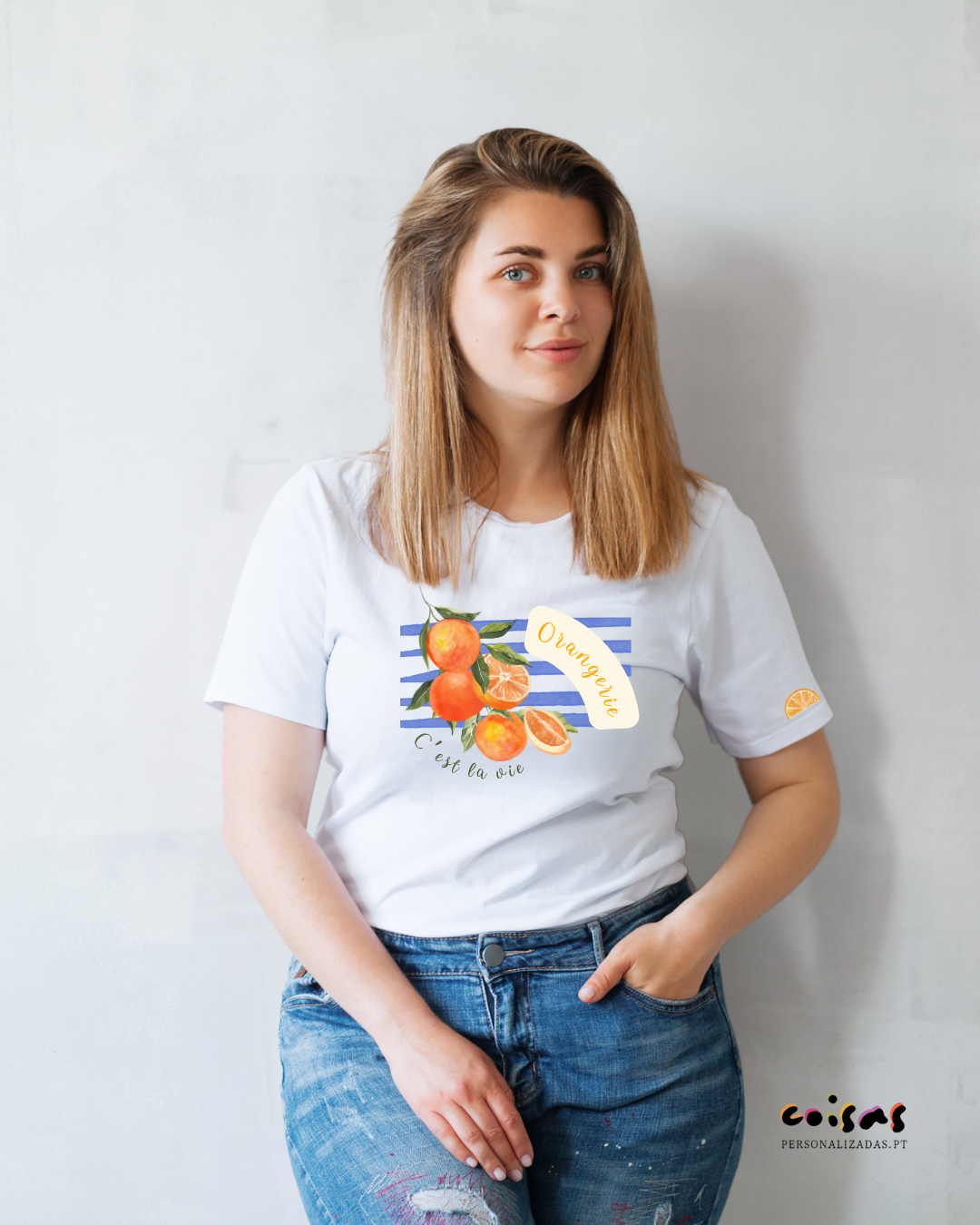 Mulher com t-shirt branca estampada de laranjas, jeans azuis, com fundo de parede clara.