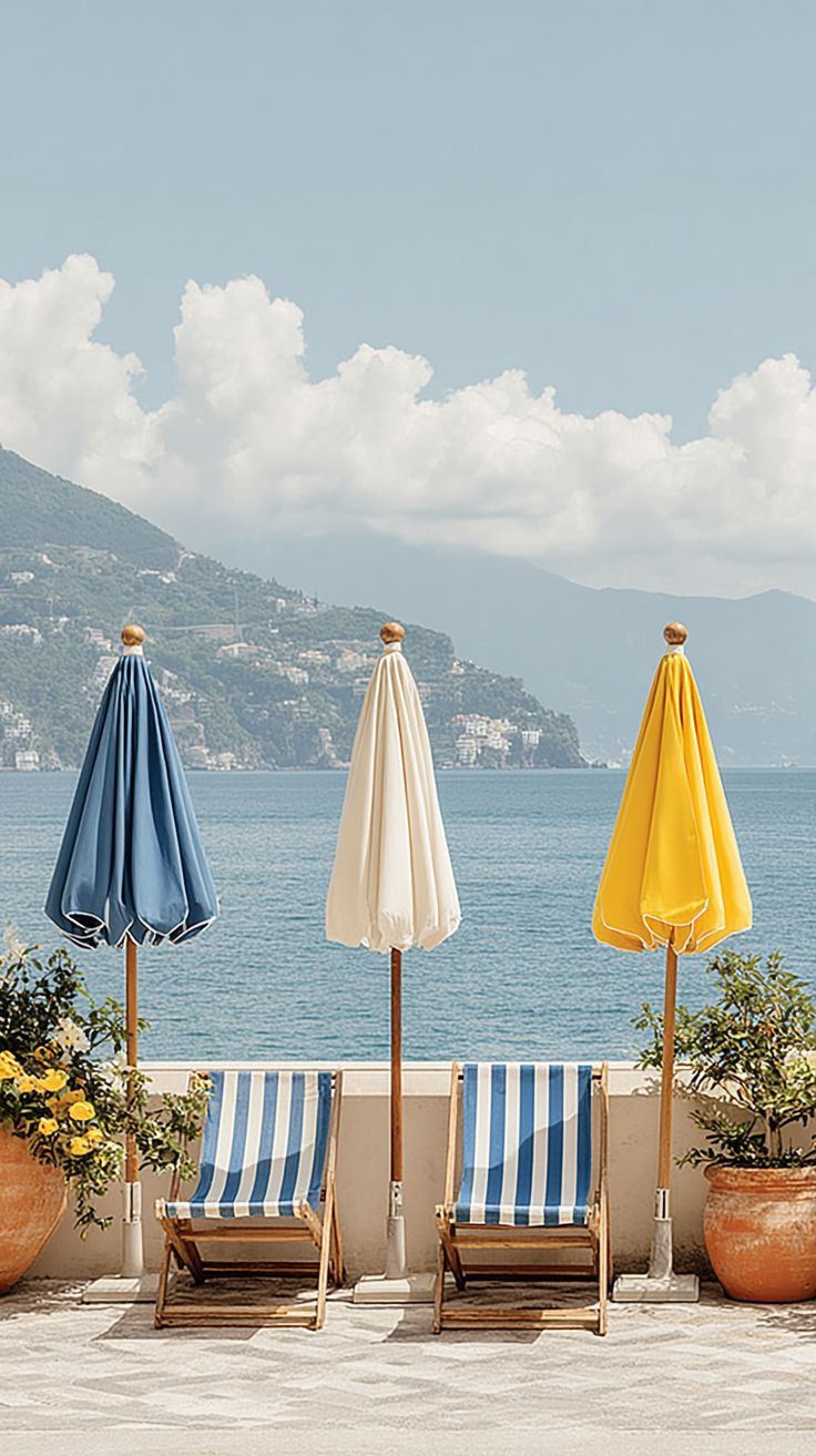 Três espreguiçadeiras às riscas azuis e brancas com três guarda-sóis fechados azul, branco e amarelo junto ao mar