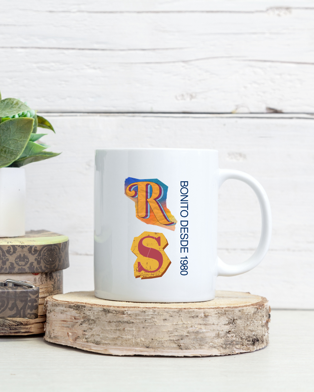 Caneca branca com letras coloridas R S e texto BONITO DESDE 1980 ao lado sobre base de madeira