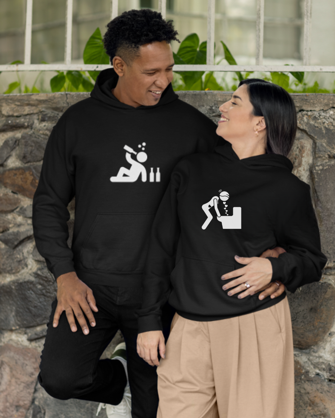 Casacos pretos com capuz com desenhos brancos de pessoa a beber e pessoa a vomitar em sanita, usados por casal junto a parede de pedra e plantas.