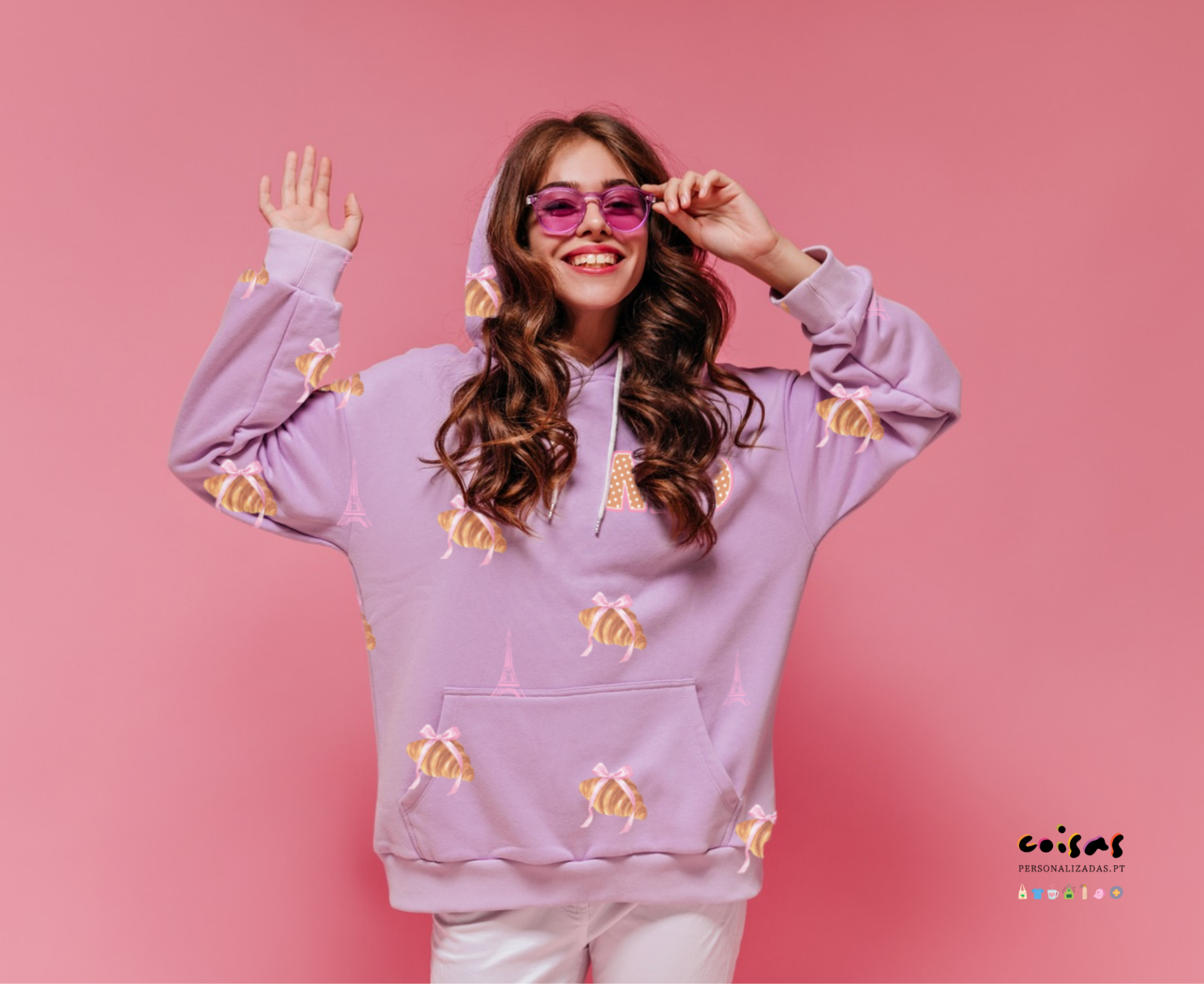 Mulher sorridente com sweatshirt lilás estampado de abelhas amarelas e torres Eiffel, usando óculos sol rosa, fundo rosa
