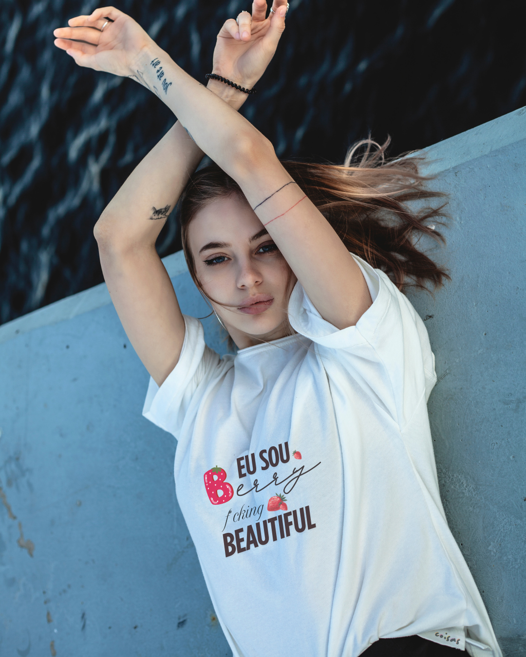 Modelo jovem com t-shirt branca estampada, posando junto a uma parede cinza e um corpo de água