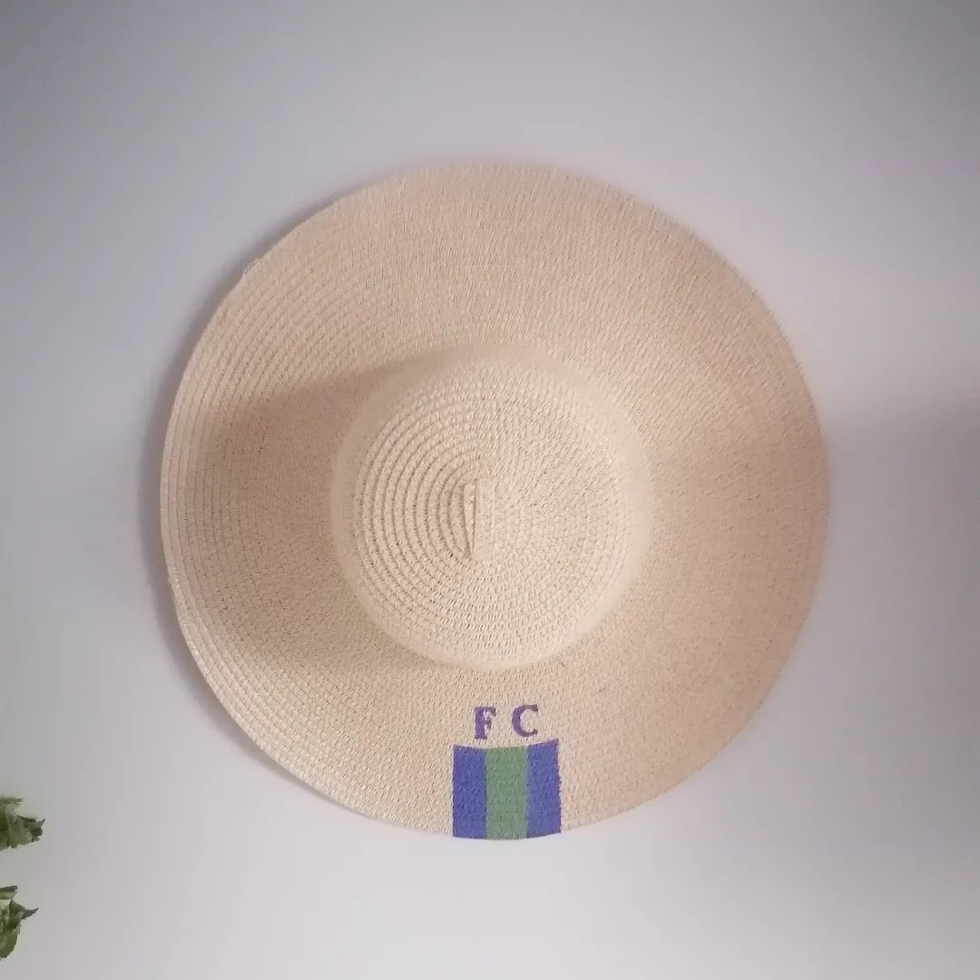 Chapéu de palha bege com letras FC e faixa verde e azul pendurado na parede