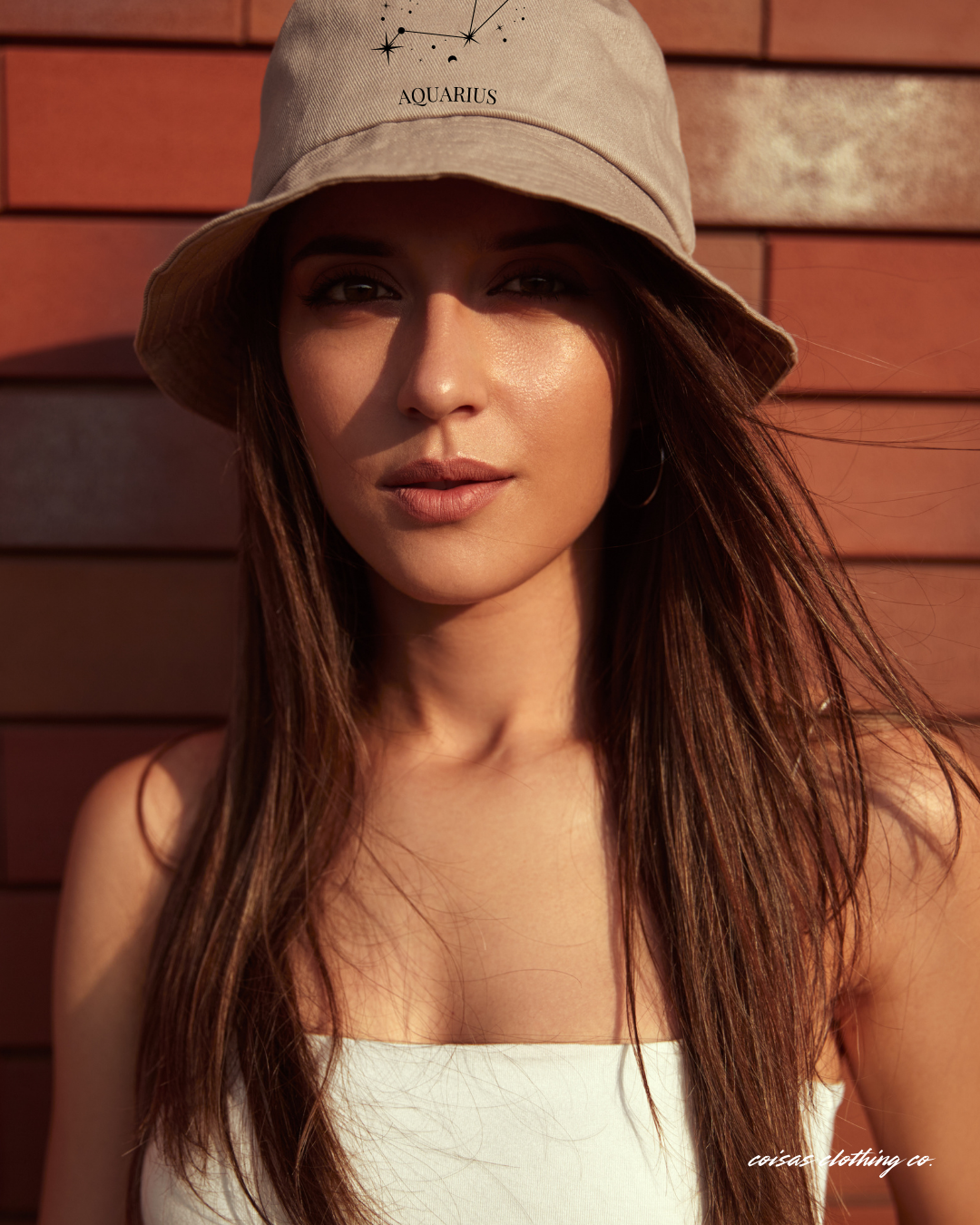 Mulher com chapéu bege claro com constelação e texto AQUARIUS em fundo de parede de madeira vermelha