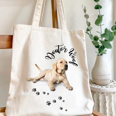 Saco de tecido cru com imagem de cão e texto 'Doutor Woof', pendurado em cadeira de madeira, com vaso branco e planta ao lado.