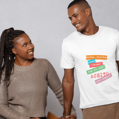 Casal sorridente de mãos dadas, homem com t-shirt branca com texto em português e mulher com camisola castanha.