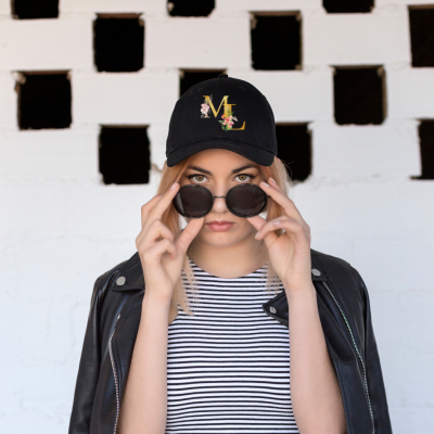 Mulher com boné preto com letra M e flor, óculos escuros, casaco de pele negro e camisola às riscas em fundo de parede branca