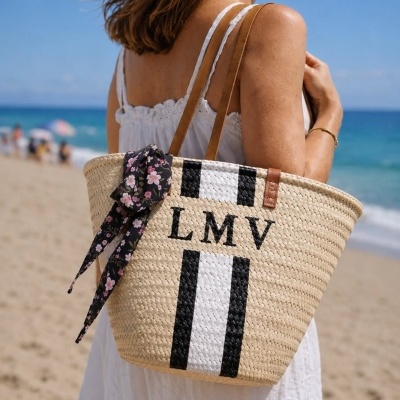Bolsa de palha com alças castanhas, listras a preto e branco e letras LMV, adornada com lenço floral, usada por mulher vestida de branco na praia
