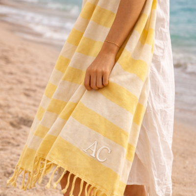 Manta de praia às riscas amarelas e brancas com franjas e monograma AC, segurada por pessoa junto ao mar.
