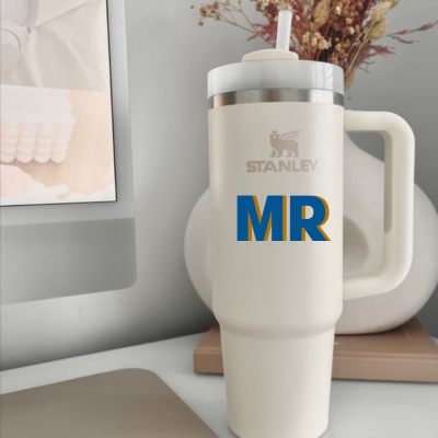 Copo térmico Stanley branco com alça grande e texto 'STANLEY' e 'MR' em azul e amarelo numa mesa com teclado e monitor