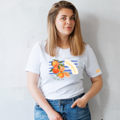 Mulher com t-shirt branca estampada de laranjas, jeans azuis, com fundo de parede clara.