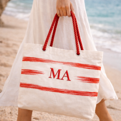 Saco de praia branco com riscas e alças vermelhas na mão de uma pessoa numa praia
