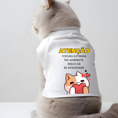 Gato cinzento com t-shirt branca e texto colorido vestindo-se sentado em superfície branca.