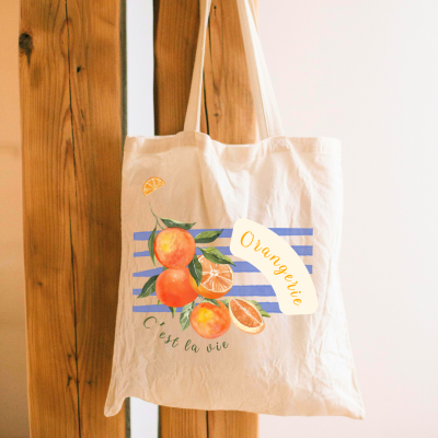 Saco de pano bege com estampado de laranjas e texto, pendurado em madeira